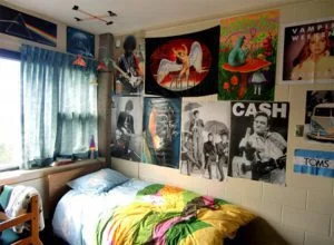 A cozy bedroom with light-colored walls decorated with various music and art posters, including images of musicians, a unicorn, and colorful abstract designs. The single bed has a vibrant yellow and green comforter, and is positioned next to a window with teal curtains, letting in natural light.