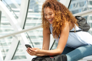 Smiling young woman with curly hair sitting in a modern architectural space, looking at her smartphone while resting her arm on a suitcase and wearing a small black backpack.
